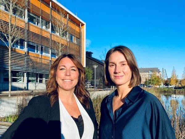 Två kvinnor från står framför en byggnad, båda ler mot kameran.
