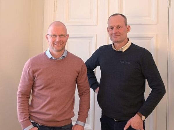 Två män poserar framför vit vägg, ena i rosa tröja och andra i svart, båda ler.