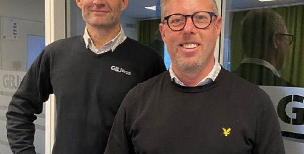 Två män i tröjor ler inomhus framför GBJ-skylt.