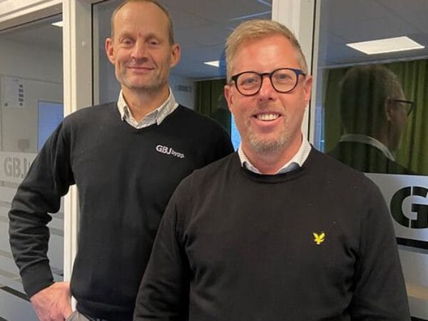 Två män i tröjor ler inomhus framför GBJ-skylt.