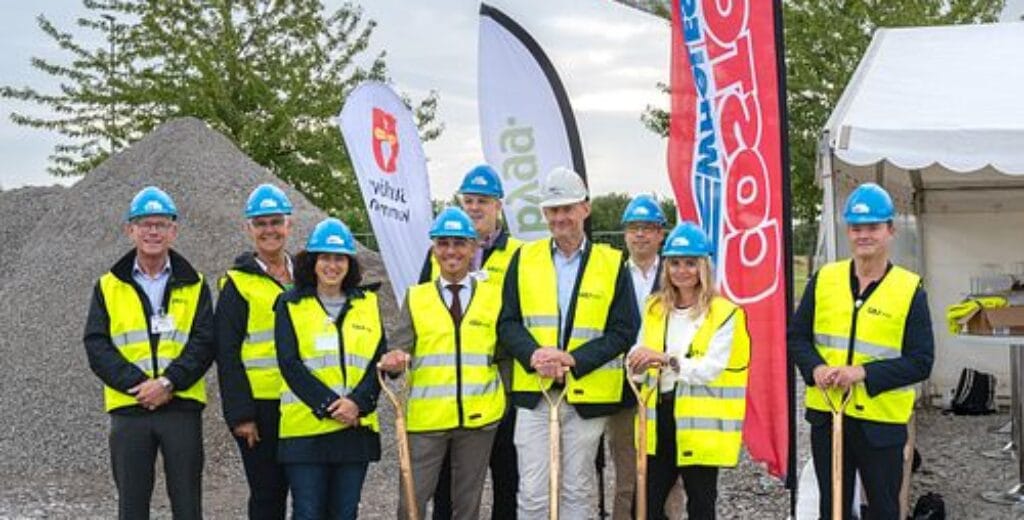 Grupp med bygghjälmar och spadar framför byggarbetsplats med flaggor.