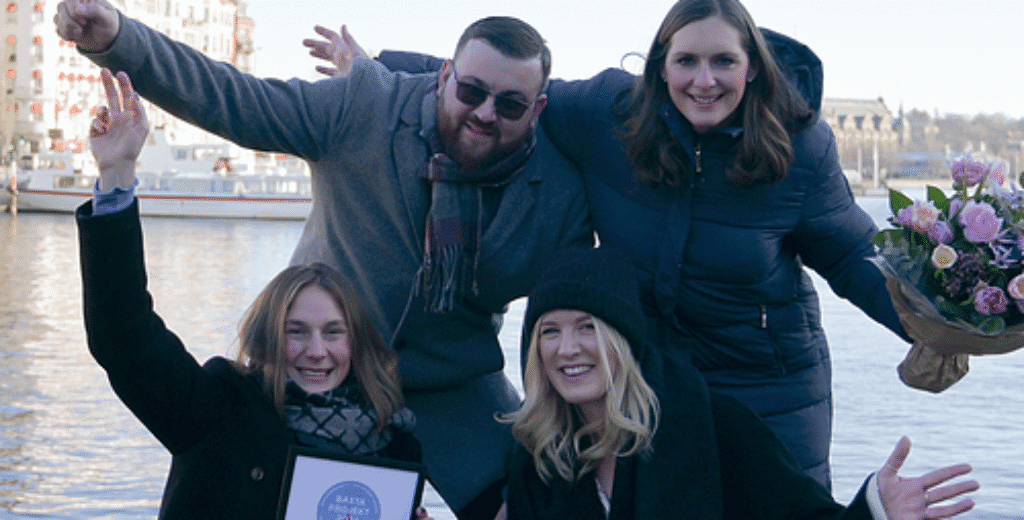 Fyra glada personer firar med diplom vid sjö, vinterklädda.