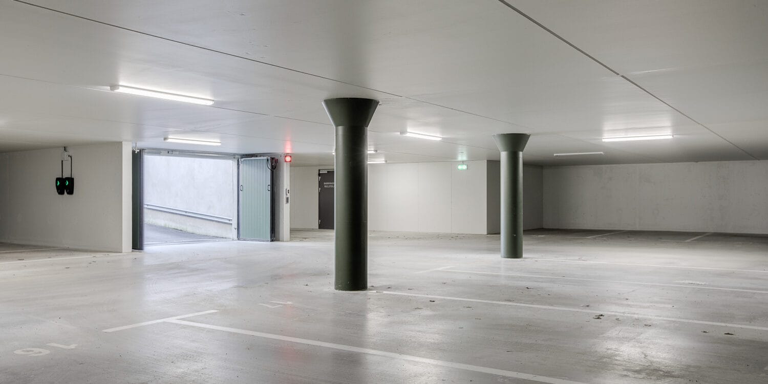 GBJ bostad har utvecklat ett ljust och rymligt parkeringsgarage med pelarstöd och elbilsladdning, där infarten syns öppen mot en betongramp som leder till gatuplan i BRF Helgablick.
