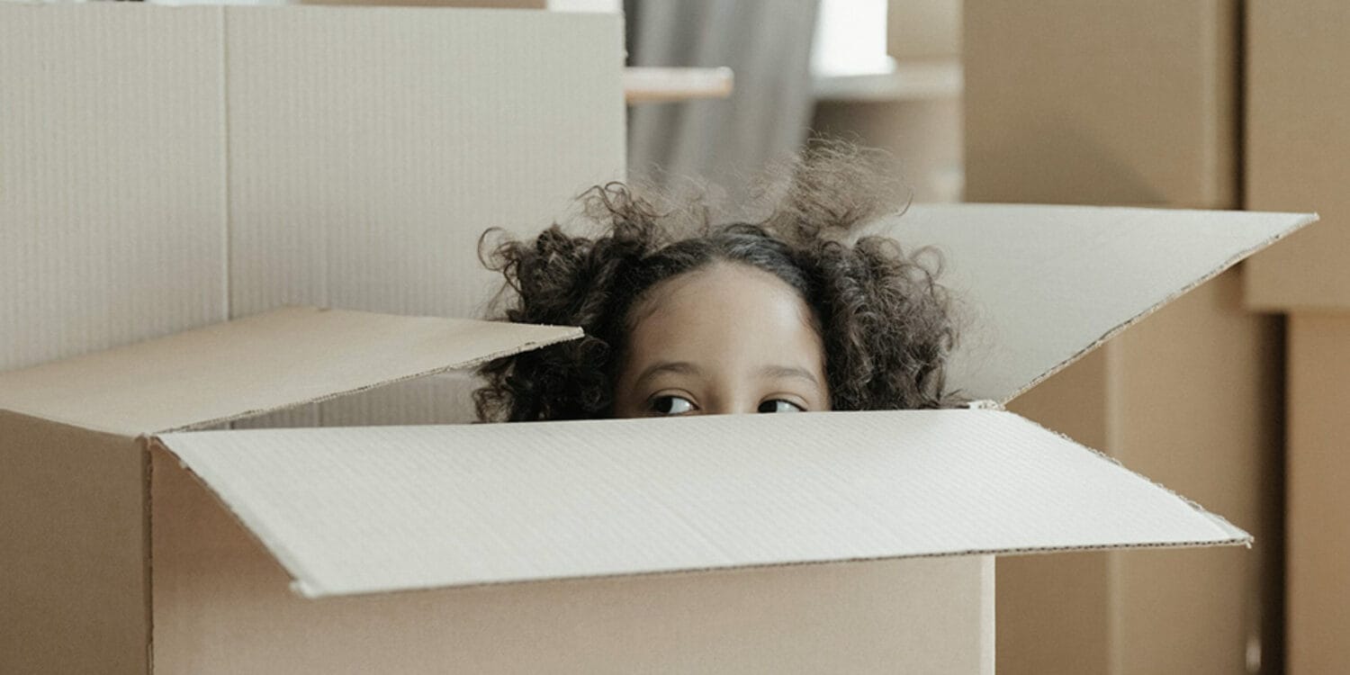 Ett barn tittar upp ur en öppen kartong bland flera flyttlådor – illustrerar familjens nya kapitel i ett hem från GBJ bostad.