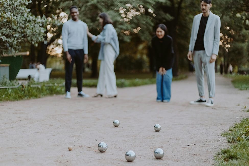 Fyra personer spelar boule i en park. Tre klot ligger på grusbanan i förgrunden medan spelarna står i bakgrunden bland grönskande träd, i en avslappnad och social atmosfär