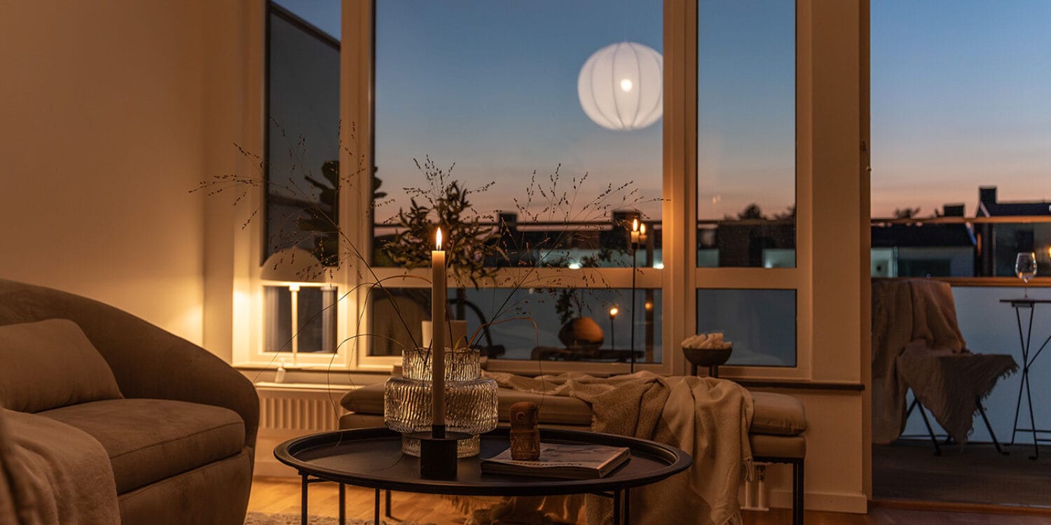 Vardagsrum i bostadsrätt i Hemmeslöv, Båstad med stämningsfull belysning och utsikt mot inglasad balkong i kvällsljus.