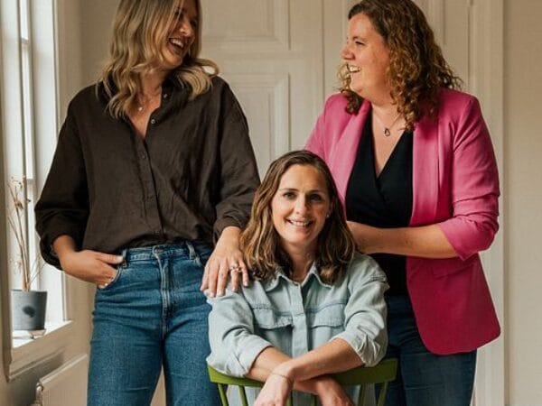 Tre kvinnor i olika åldrar skrattar tillsammans i hemmiljö – en sittande, två stående.