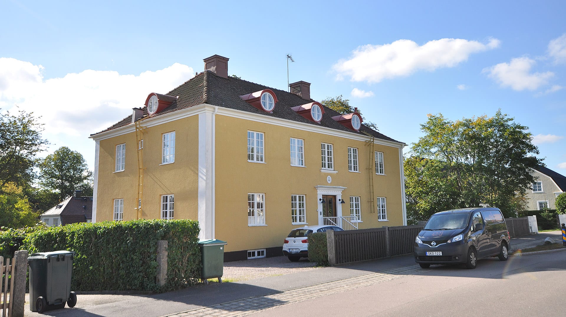 Charmigt äldre hyreshus i gult med röd takpanna, beläget i ett lugnt bostadsområde med goda parkeringsmöjligheter.