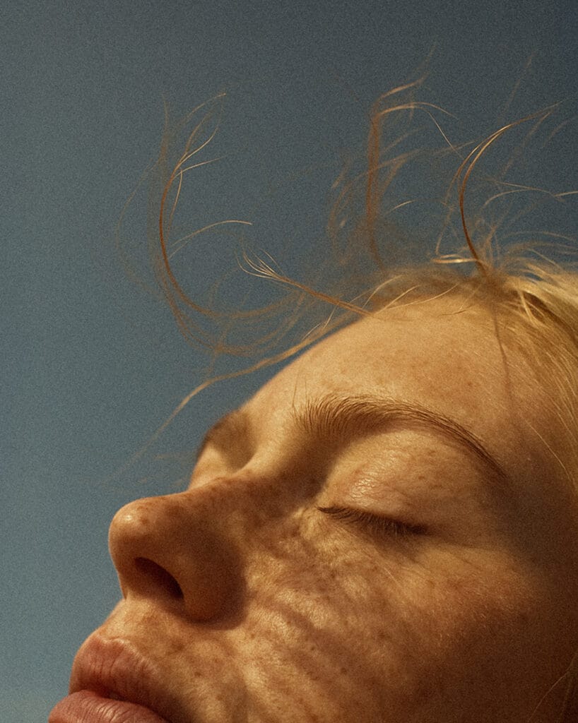 Ett närbildsporträtt av en kvinna med fräknar och slutna ögon fångar en stilla stund i solen, där vinden leker i håret – en symbol för livskvalitet i Ängsrum, ett bostadsprojekt från GBJ bostad.