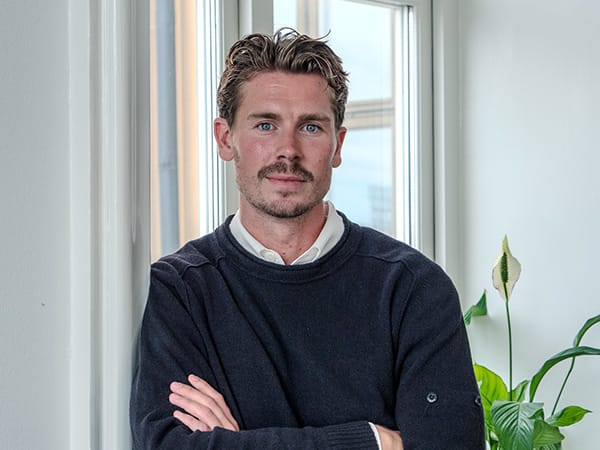 En man står med armarna i kors framför ett fönster med växter, i ett porträtt som representerar en del av teamet bakom GBJ bostads bostadsutveckling.