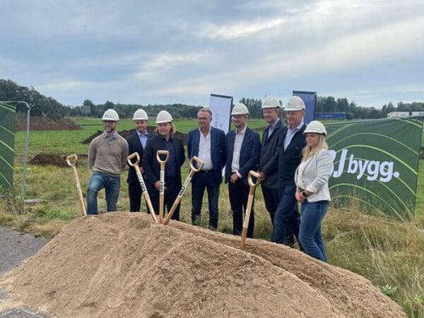 Sju personer med hjälmar vid grön byggskylt, poserar på grushög vid spadtagsceremoni.