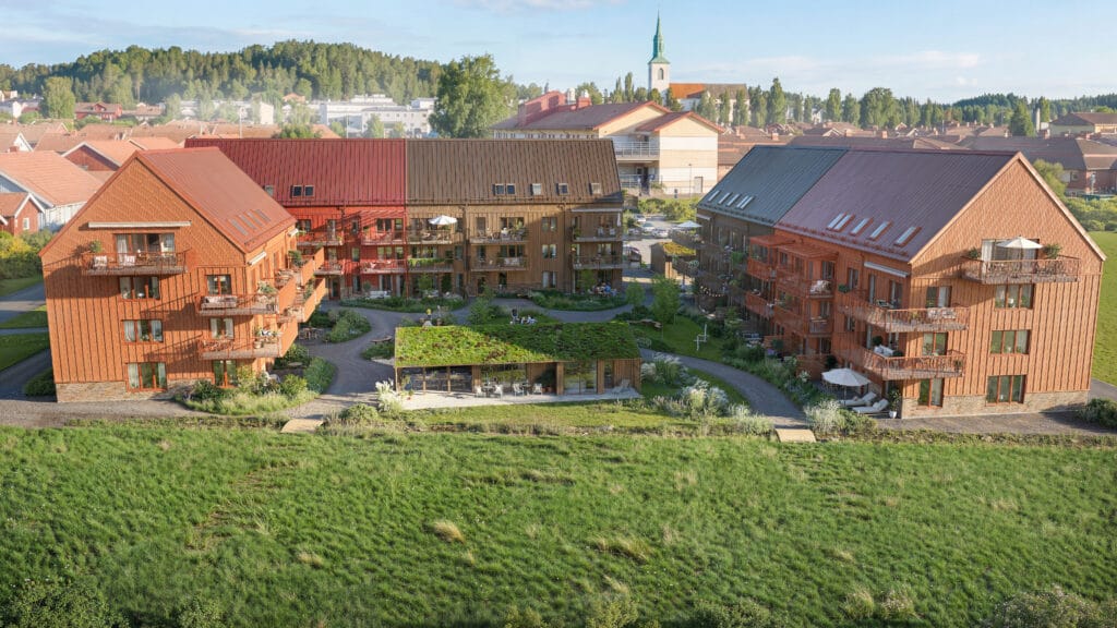 Översikt Uppdaterad.jpg visar flerbostadshusen i projekt Ängsrum, utvecklat av GBJ bostad, där tre träklädda byggnader med röda och mörka tak omger en grön innergård med gemensamhetsyta och sedumtak. Balkonger och uteplatser vetter mot slingrande gångar i en småskalig stadsmiljö med kyrktorn och villabebyggelse i bakgrunden, vilket skapar en trygg och naturnära boendemiljö.