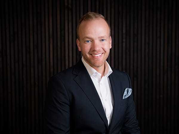 Gustav Johansson från Nya Kvadrat, klädd i mörk kostym, poserar leende mot en mörk träpanel – samarbetande mäklare för GBJ bostads projekt i Ytterby.