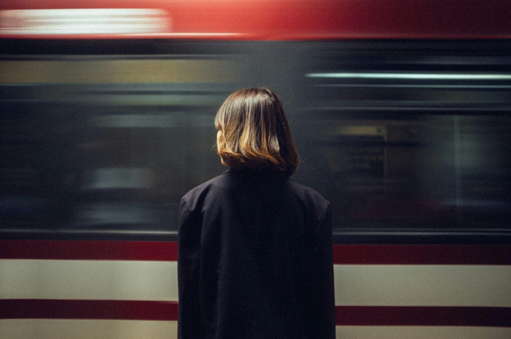 En person står på en pendeltågsplattform medan ett tåg passerar i bakgrunden, suddigt av rörelse, i en urban miljö som speglar smidig pendling nära bostäder utvecklade av GBJ bostad i Ängsrum Ytterby.